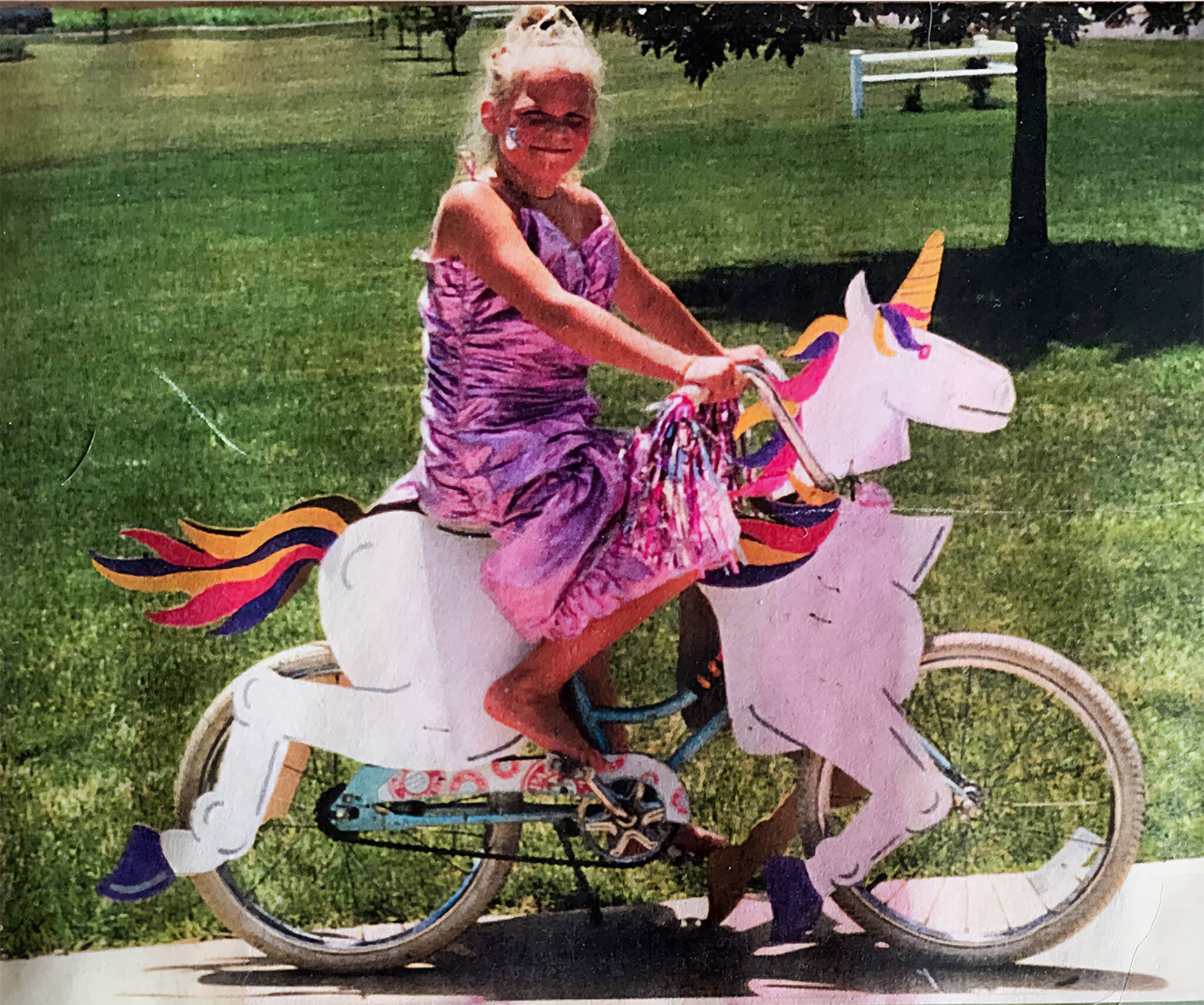 Curt's daughter as a child, a blonde girl wearing a pink metallic dress, riding a bicycle outfitted with a running unicorn cutout.
