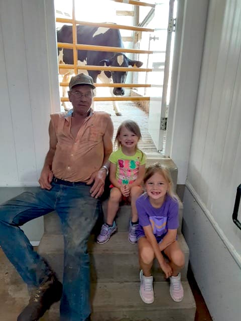 Older dairy farmers with two blonde grand daughters and a dairy cow in the background.